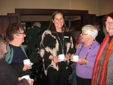 A group of women, most holding cups and saucers chat in a crowded room.