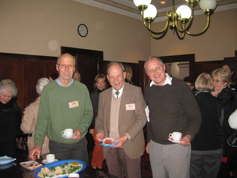 Three men in a crowded room, standing near a table with biscuits and sandwiches, smile for the camera. Two of the men are wearing name badges. Two are holding cups and saucers and the other has a plate with food.