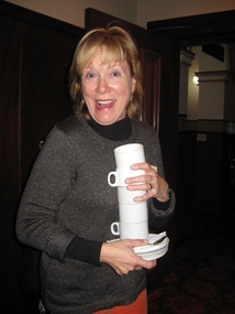 A woman acts surprised when she is caught by the camera carrying a stack of cups and saucers.