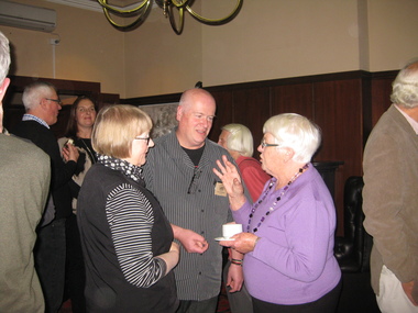 Two women and a man chat in a crowded room. the woman on the right is holding a cup and saucer.