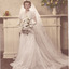A woman in a white wedding dress, holding a bouquet of white flowers stands in front of a fireplace with a white surround and mantlepiece. A vase of colourful flowers stand on the right end of the mantle.