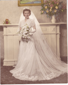 A woman in a white wedding dress, holding a bouquet of white flowers stands in front of a fireplace with a white surround and mantlepiece. A vase of colourful flowers stand on the right end of the mantle.