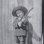 A small boy standing holding rifle, he is wearing a jacket and short pants.  