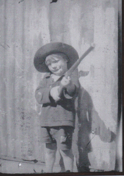 A small boy standing holding rifle, he is wearing a jacket and short pants.  