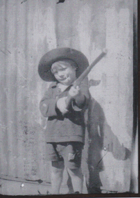 A small boy standing holding rifle, he is wearing a jacket and short pants.  