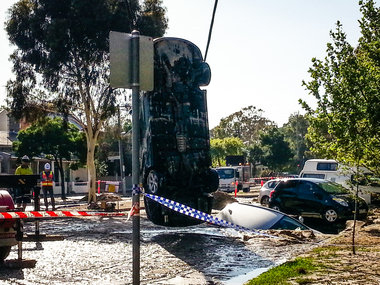 car being lifted from sink hole, another car is still in the sink hole.  2 workmen are watching, other cars are seen parked on street.
