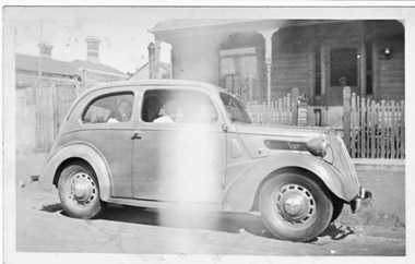 Some people sitting in a car outside a weatherboard house.