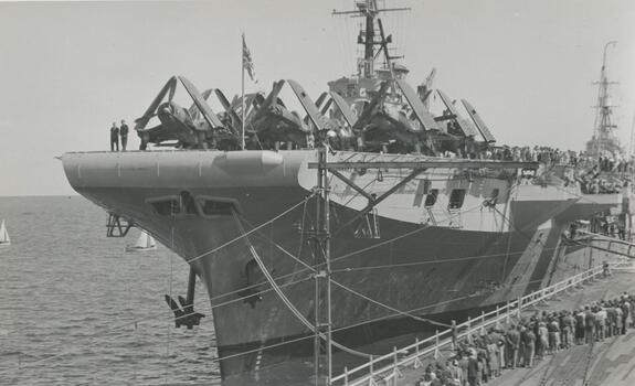 Photo shows the stern of the British aircraft carrier HMS Glory at Station Pier, Jan 1946. You can also see the aircraft that were used on this ship and the crowds lining up to inspect this ship.