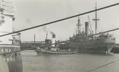 Photo shows the tug bug 'Marimba' perpendicular, stern first, to a cargo ship viewed through the mooring ropes of another ship.