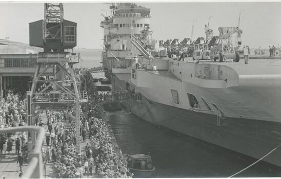 Photo shows crowds lining up to board the British aircraft carrier HMS Indefatigable at Station Pier, Jan 1946. You can see machinery lined up on the side of the aircraft carrier. 