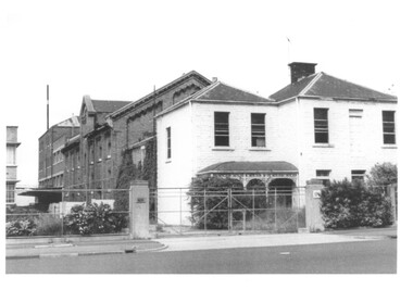 Several buildings including a two storey house behind cyclone fencing and double gates.