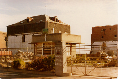 Partly demolished factory buildings behind a cyclone wire fence.