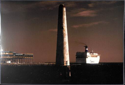 A lighthouse-like structure dominates the centre foreground. On the right a white ship is sailing away from a pier seen on the left.