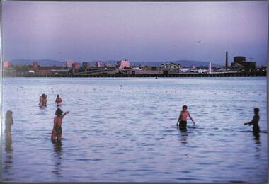 Several people a wading in the sea with the buildings on the foreshore in the background.