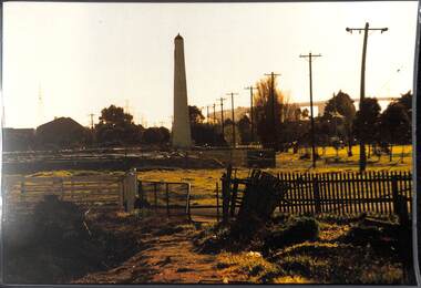 Looking from vacant land within a dilapidated wooden fence towards a lighthouse-like structure with a large road bridge in the far distance.