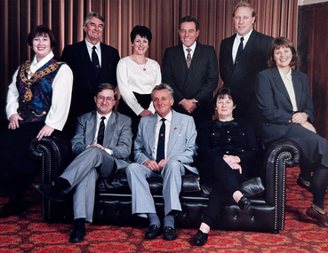 Nine people, 6 men and 3 women, posing for the camera. Two men and a woman are sitting on a black couch, two women are sitting on the arms of the couch (one on each one) and three men and a woman are standing behind the couch.