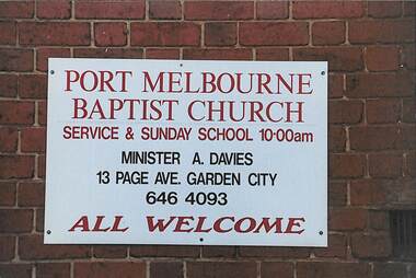 White sign with red and black writing attached to a brick wall.