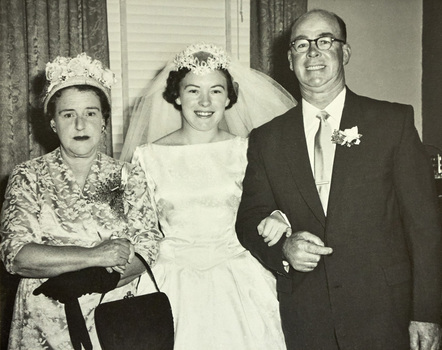 A bride with a woman wearing a dress, hat and one glove carrying a handbag and the other glove on her right and a man in a suit on her left