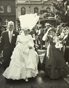 A bride with her veil blowing in the wind walks towards the camera flanked by a man in a suit and a bridesmaid