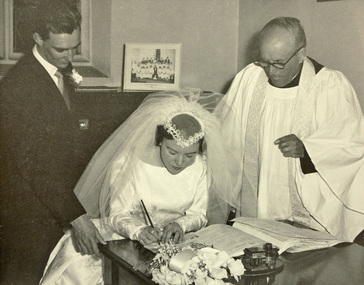 A bride signs the wedding register as a man in a suit and a clergyman look on