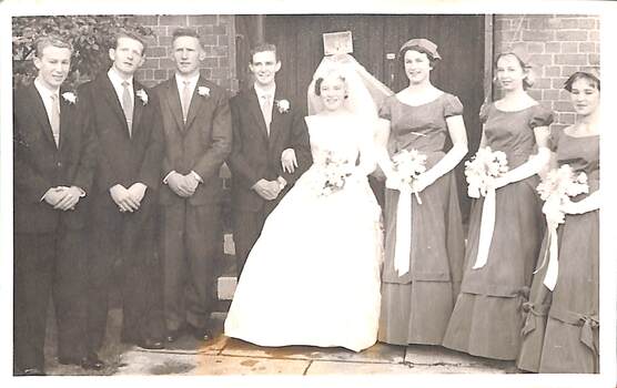 Four men in suits, a bride in wedding dress and three bridesmaids stand outside a doorway