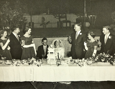 Bride and groom sitting at a wedding table behind a multi-tier wedding cake being toasted by the wedding party.