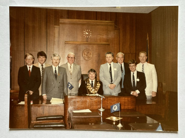 Eight men wearing suits and ties standing, four either side, of a man seated  wearing mayoral chains.