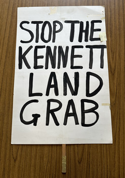 White cardboard placard attached to a wooden handle with black hand-printed slogan - Stop The  Kennett Land Grab