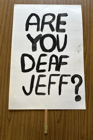 White cardboard placard attached to a wooden handle with black hand-printed slogan - Are You Deaf Jeff?