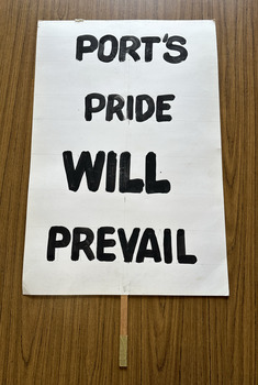 White cardboard placard attached to a wooden handle with black hand-printed slogan - Port's Pride Will Prevail
