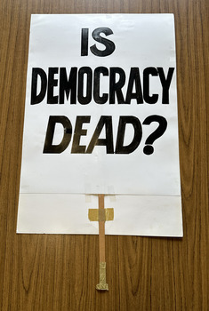 White cardboard placard attached to a wooden handle with black hand-printed slogan - Is Democracy Dead?