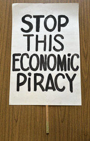 White cardboard placard attached to a wooden handle with black hand-printed slogan - Stop This Economic Piracy