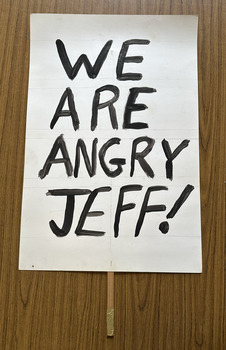 White cardboard placard attached to a wooden handle with black hand-printed slogan - We Are Angry Jeff!