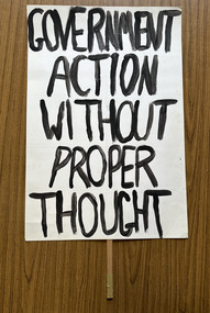 White cardboard placard attached to a wooden handle with black hand-printed slogan - Government Action Without Proper Thought