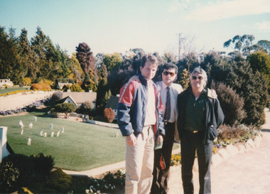 Three men posing on a sunny day in front of a cricket ground in a model village