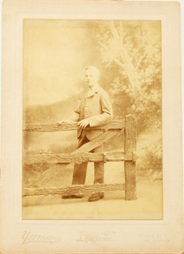 Studio photograph of boy in a suit standing behind a fence.