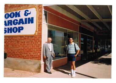 An older and young man standing under a verandah at the end of a shopping strip.