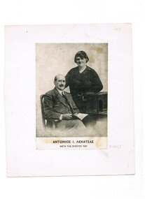 A married couple with the man seated and his wife standing next to him.