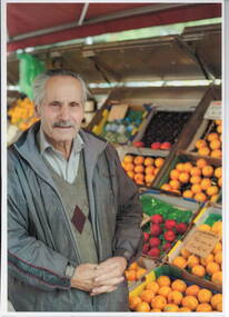 A coloured photograph of a fruit vendor standing beside his fruit barrow.