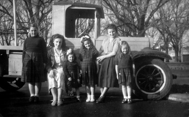 Two women and four girls standing in front of a vintage Chevrolet truck which has an open tray behind the cabin.