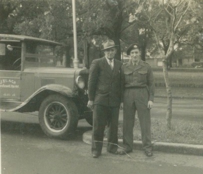 Two men, one dressed in Australian Army uniform, are standing beside a Chevrolet truck, which is parked in a suburban street.