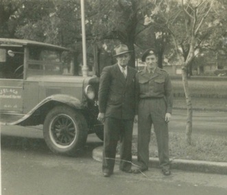 Two men, one dressed in Australian Army uniform, are standing beside a Chevrolet truck, which is parked in a suburban street.