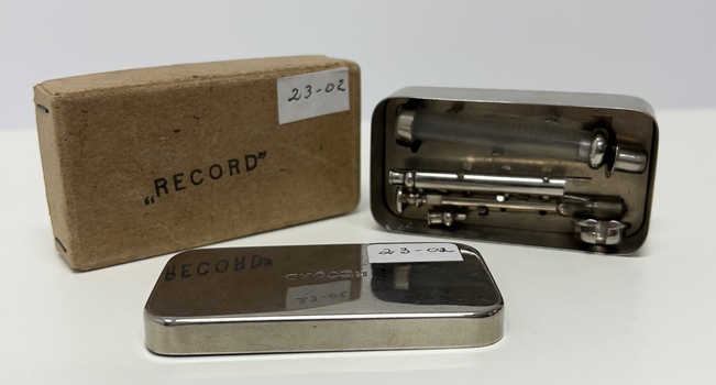 Picture shows a small cardboard box used to hold the small metal  box containing glass syringes and metal needles. Cardboard box and metal box have RECORD stamped on the lid.