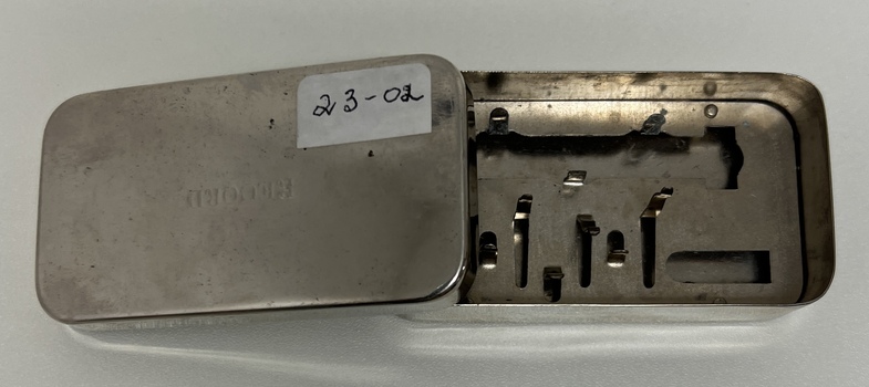 Picture shows a small metal  box containing spaces where the glass syringes and metal needles are housed. 