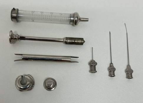 Picture of glass syringe and metal plunger along with a number or reusable needles.