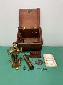 A wooden box featuring a metal microscope, a key, additional eye piece, box of glass slides and cylindrical eye piece, and lens attachment.