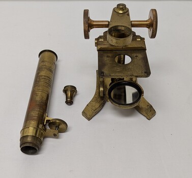 Photograph of a Brass and glass microscope made of 3 parts consisting of a Brass body, eye piece and eye piece adaptor. Photo shows 3 separate pieces.