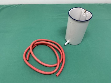 A red rubber tube lies coiled beside a white enamel canister with a blue rim, small side spout at the base.