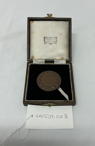 A round bronze medallion in a velvet-lined presentation box with a ribbon, displayed on a white surface with a tag labeled "202509.008.