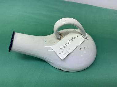 White ceramic vessel with a rounded body, arched handle, and short side spout, resting on green fabric with a handwritten ID tag.